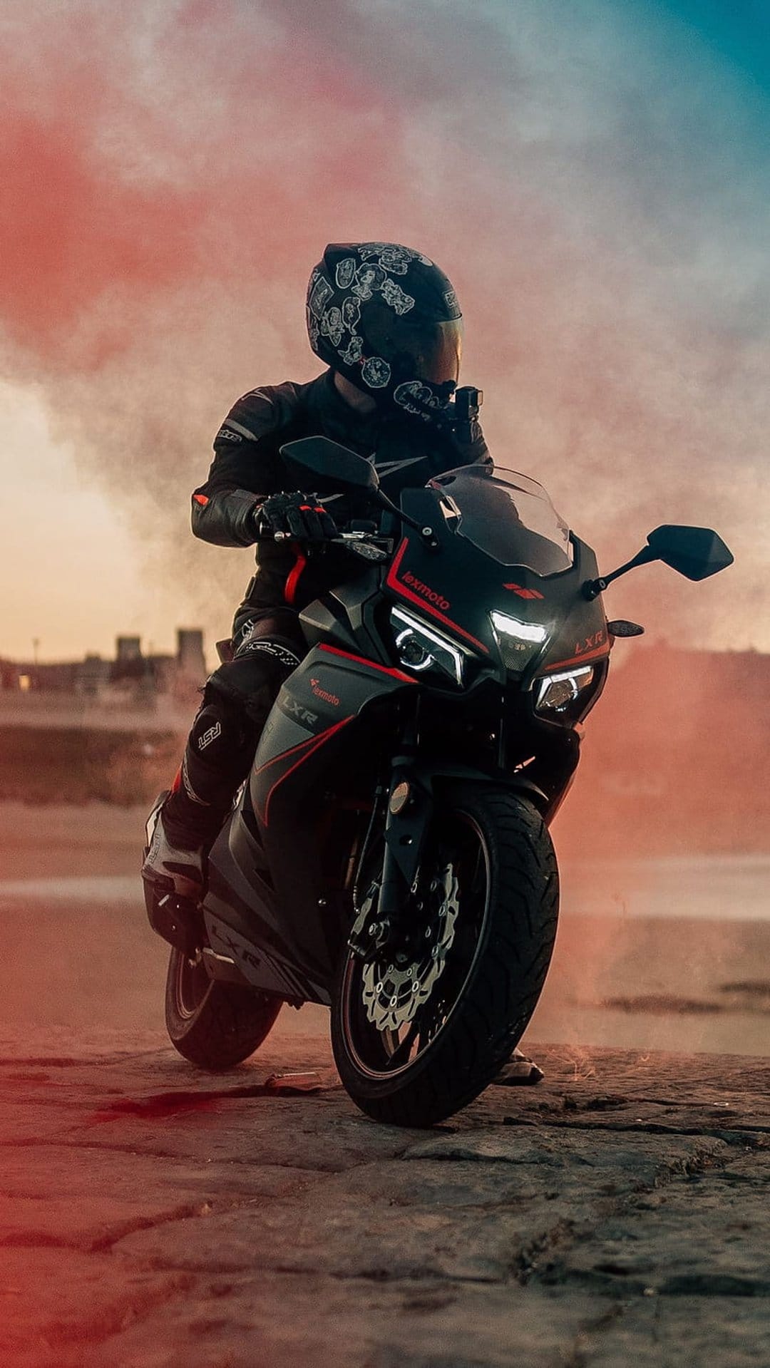 Motorcyclist in full gear on a red and black sportbike atop rocks near water during a dramatic sunset.