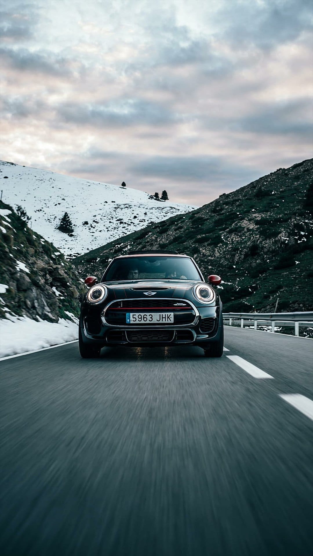 Black Mini Cooper navigating a winding asphalt road through snowy alpine peaks under a cloudy sky at dusk.