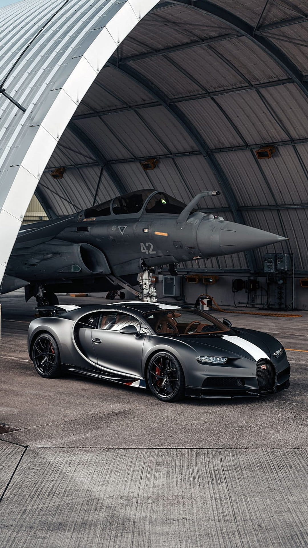 Bugatti Chiron supercar parked in a hangar with a fighter jet, featuring a white stripe and grey paint finish.
