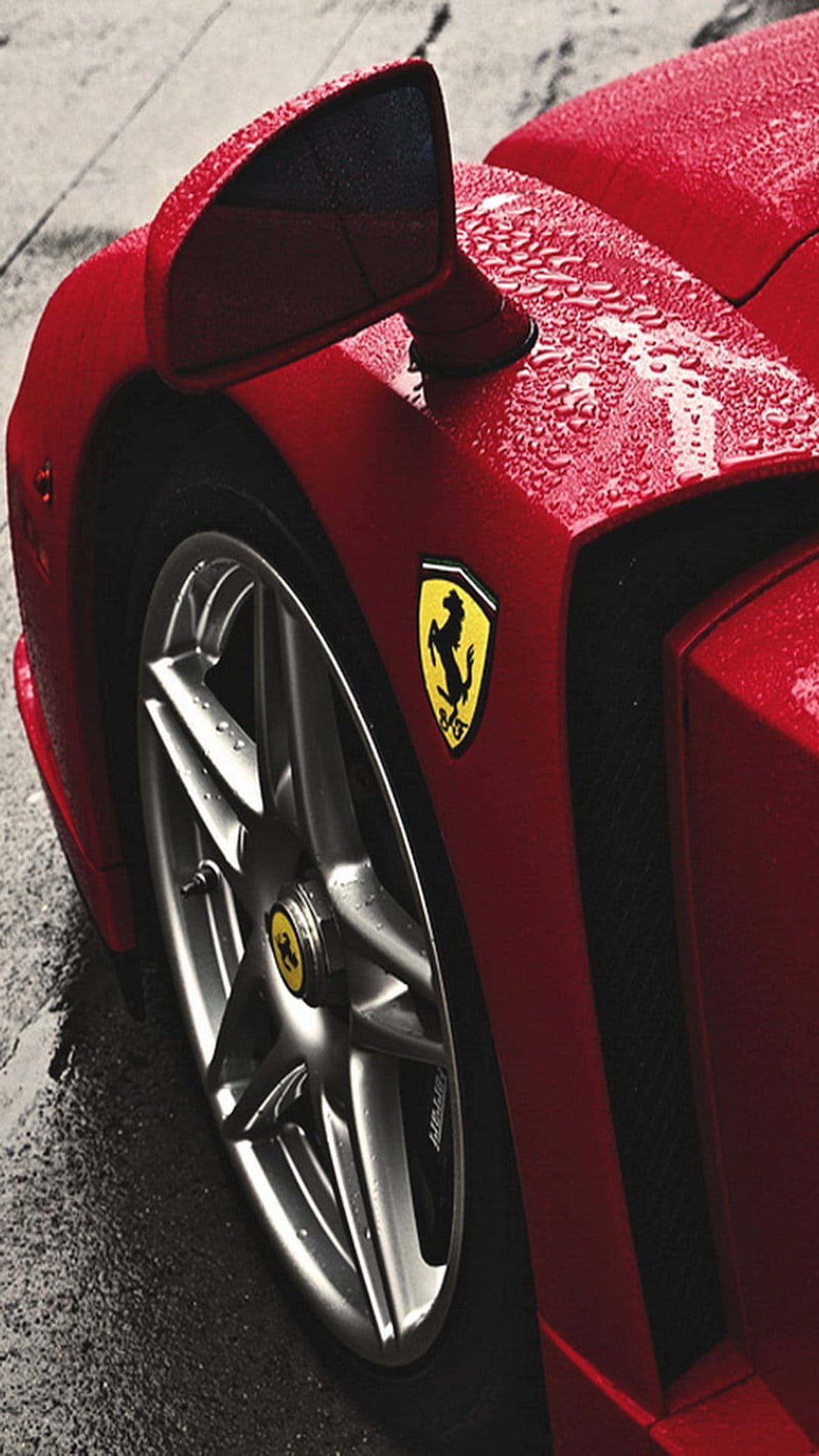 Red Ferrari fender with yellow prancing horse emblem and wet alloy wheel covered in glistening rain droplets.