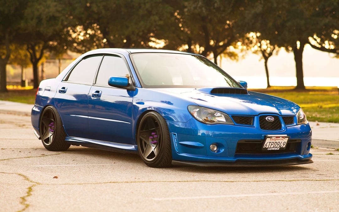 Blue Subaru WRX STI with custom wheels and lowered suspension parked at sunset with trees in the background.