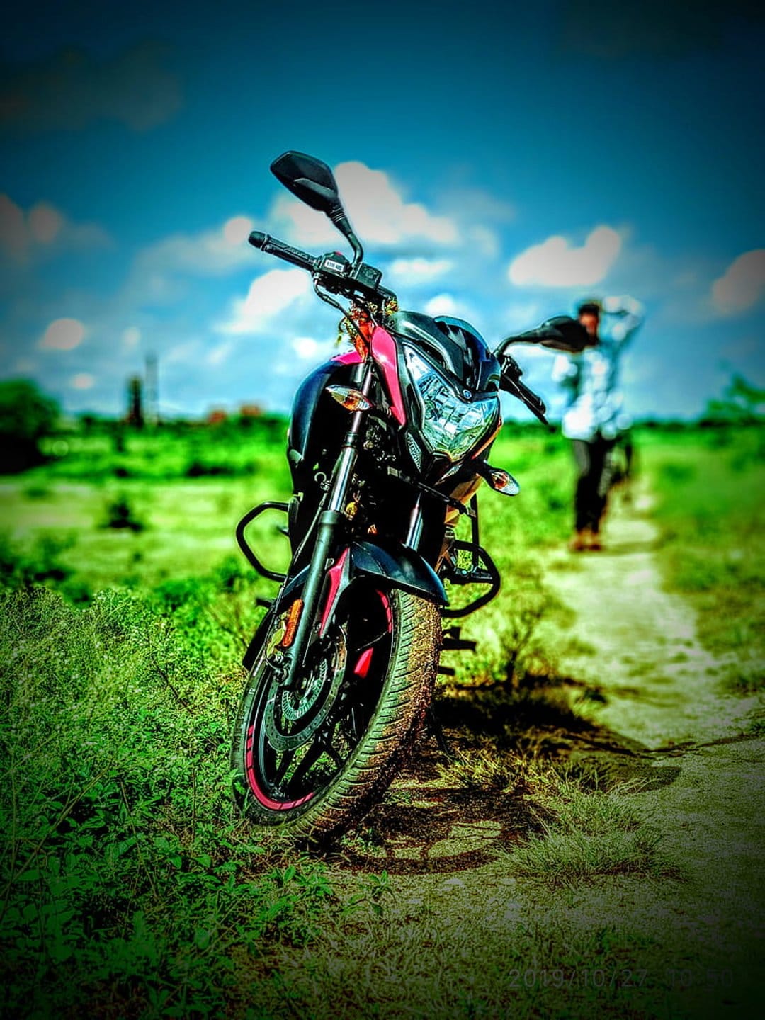 Sporty motorcycle parked on a green field with a blurred person walking away under a bright cloudy blue sky.