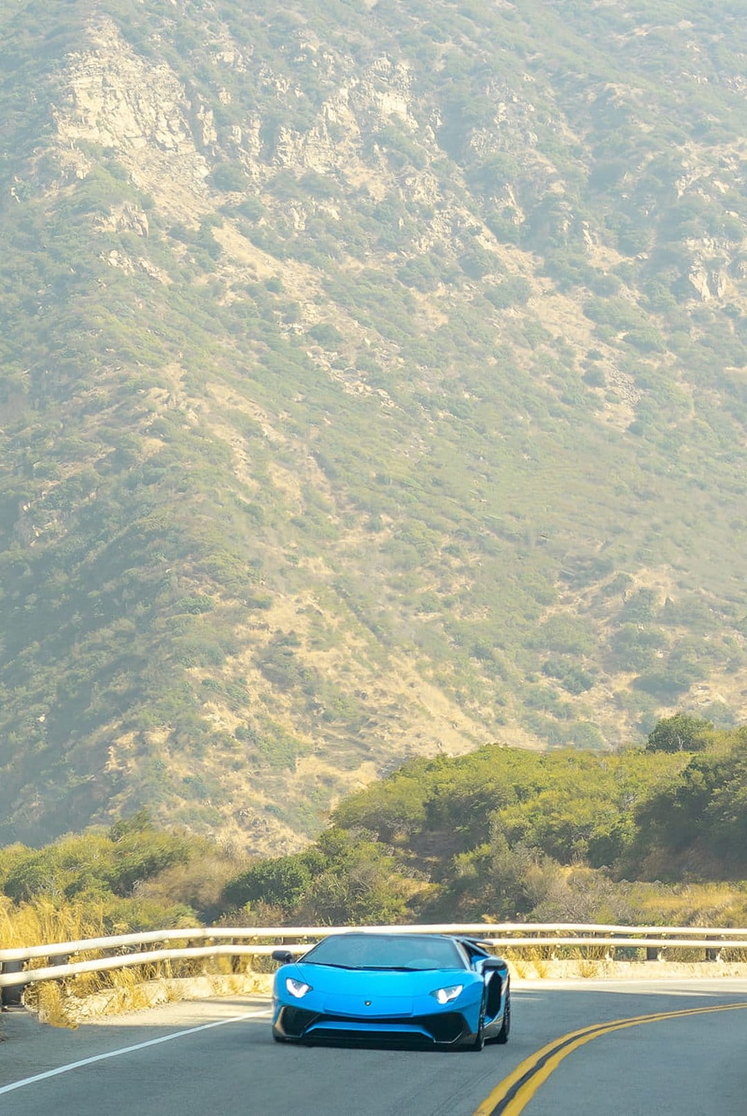 Blue Lamborghini Aventador supercar driving on a winding mountain road surrounded by green hills and sunlight.