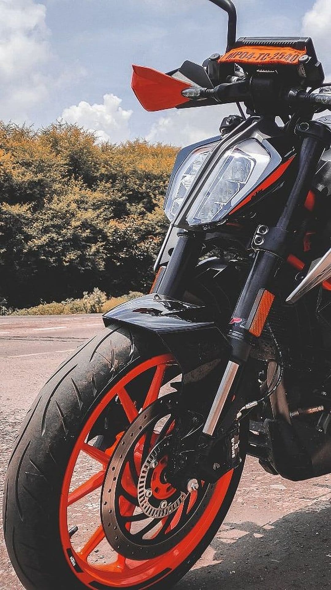 KTM motorcycle front wheel with an orange rim and headlight assembly set against green foliage and cloudy sky.