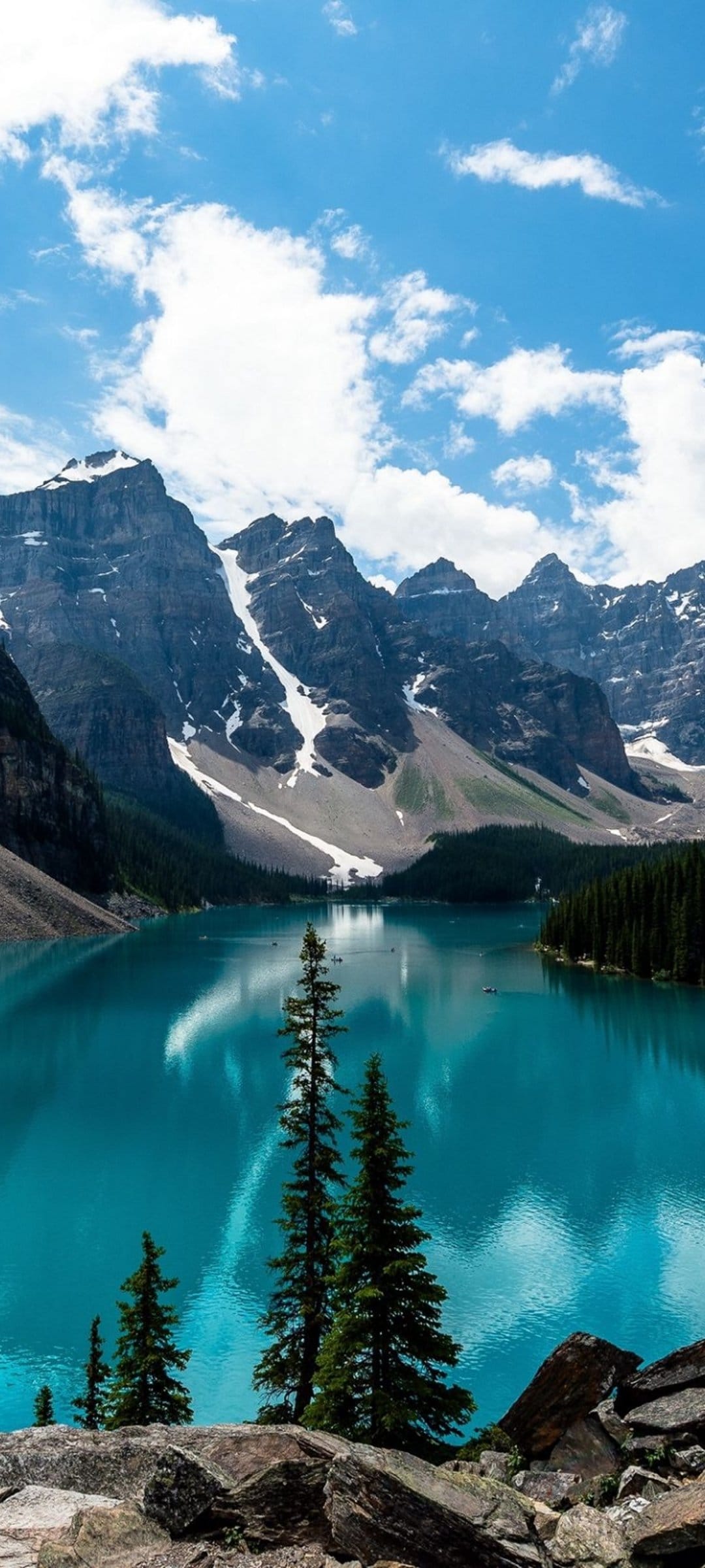 Vibrant teal lake water reflects tall snow-covered mountains and evergreen trees in a vertical wilderness scene.