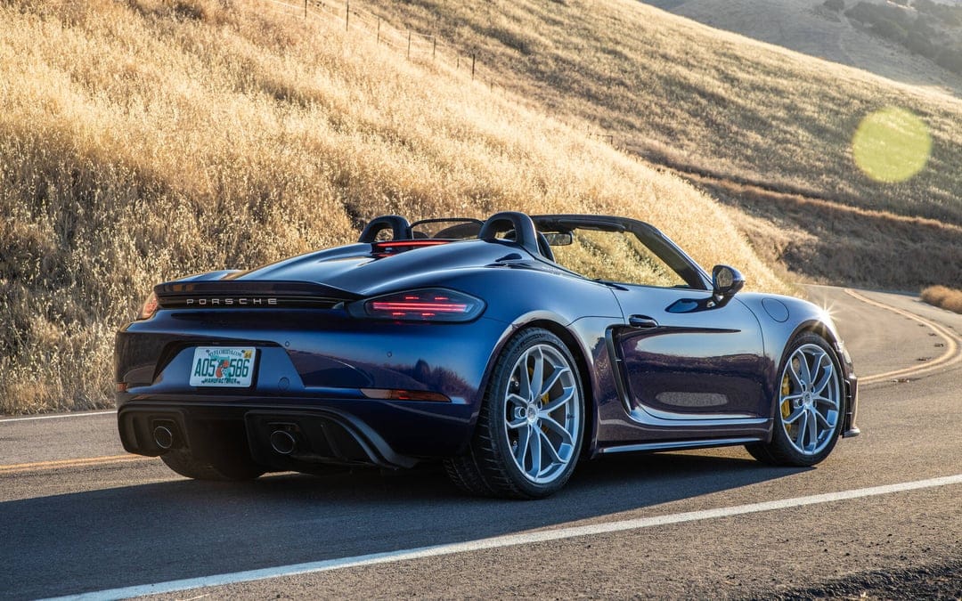 Dark blue Porsche 718 Spyder convertible on a winding mountain road with golden sunlit hills in the background.