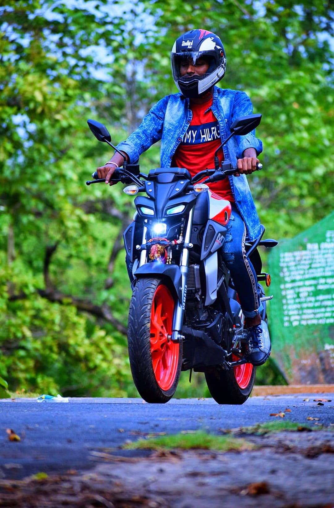 Man on a black motorcycle with orange wheels rides along a paved forest road lined with dense green foliage.