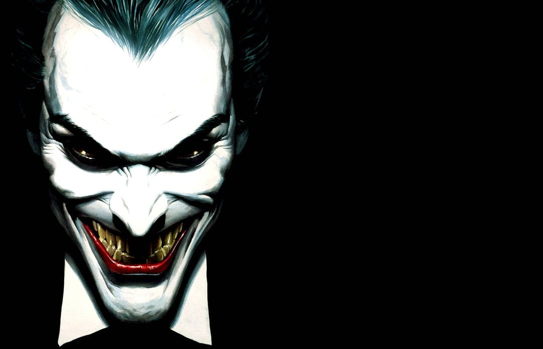 Joker with white face paint and sharp yellow teeth grins against a black background in a tight portrait view.