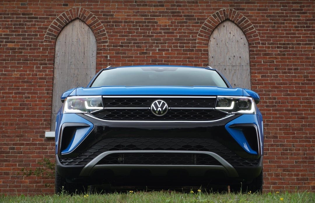 Front view of a blue Volkswagen SUV parked on green grass against a rustic red brick wall with arched windows.