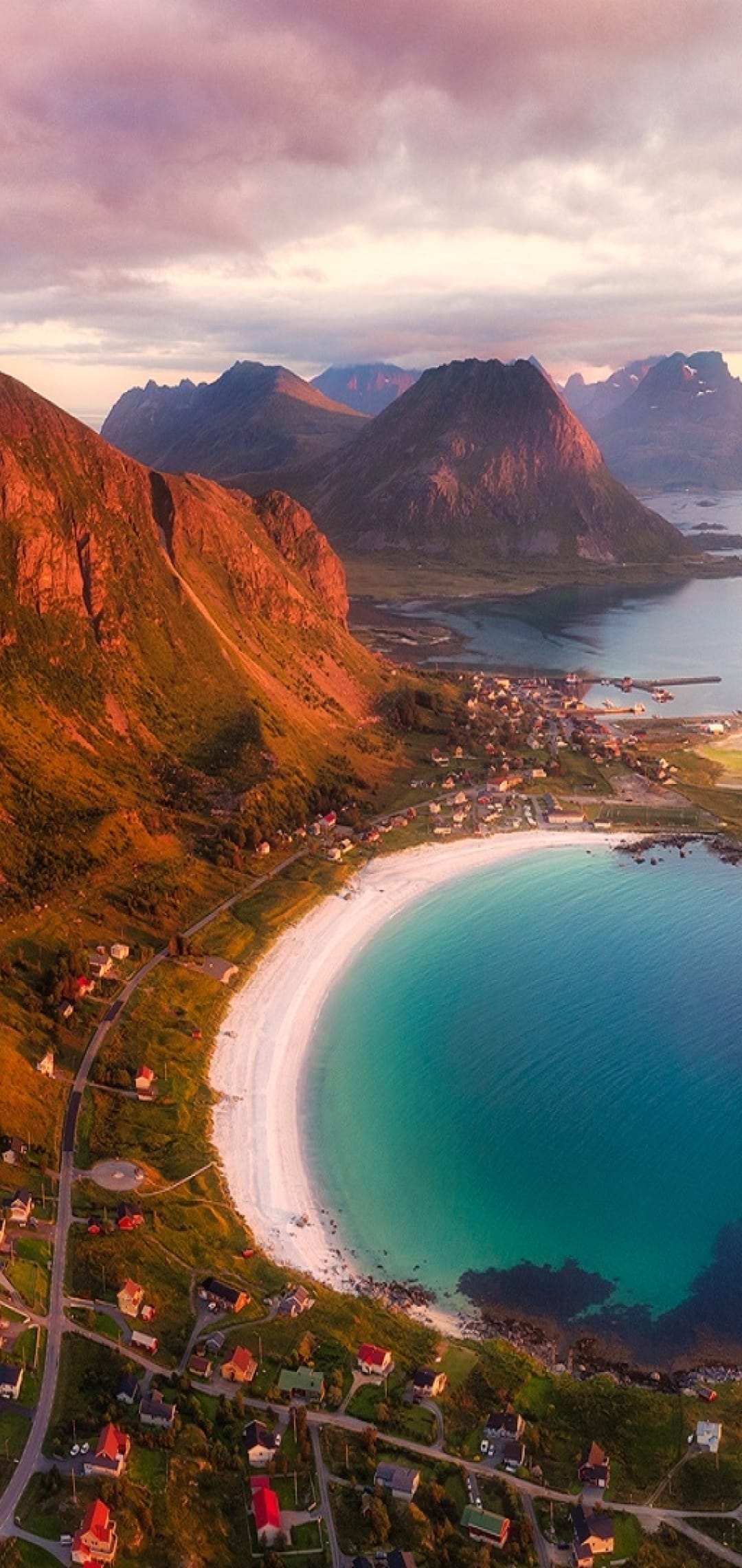 Coastal village houses along a turquoise bay and white beach beneath jagged mountains during a pastel sunset.