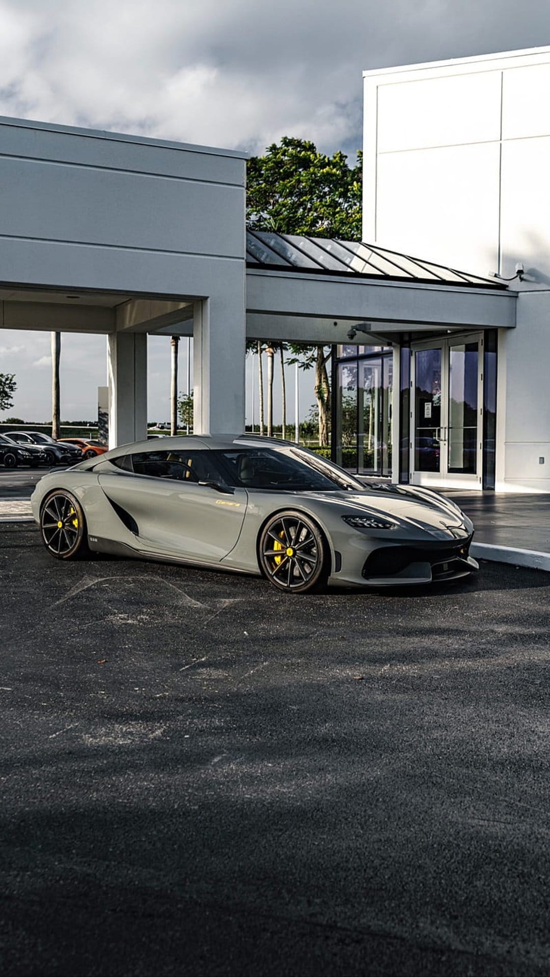 Grey sports car with yellow brake calipers parked near a glass building with cloudy sky reflections on the hood.