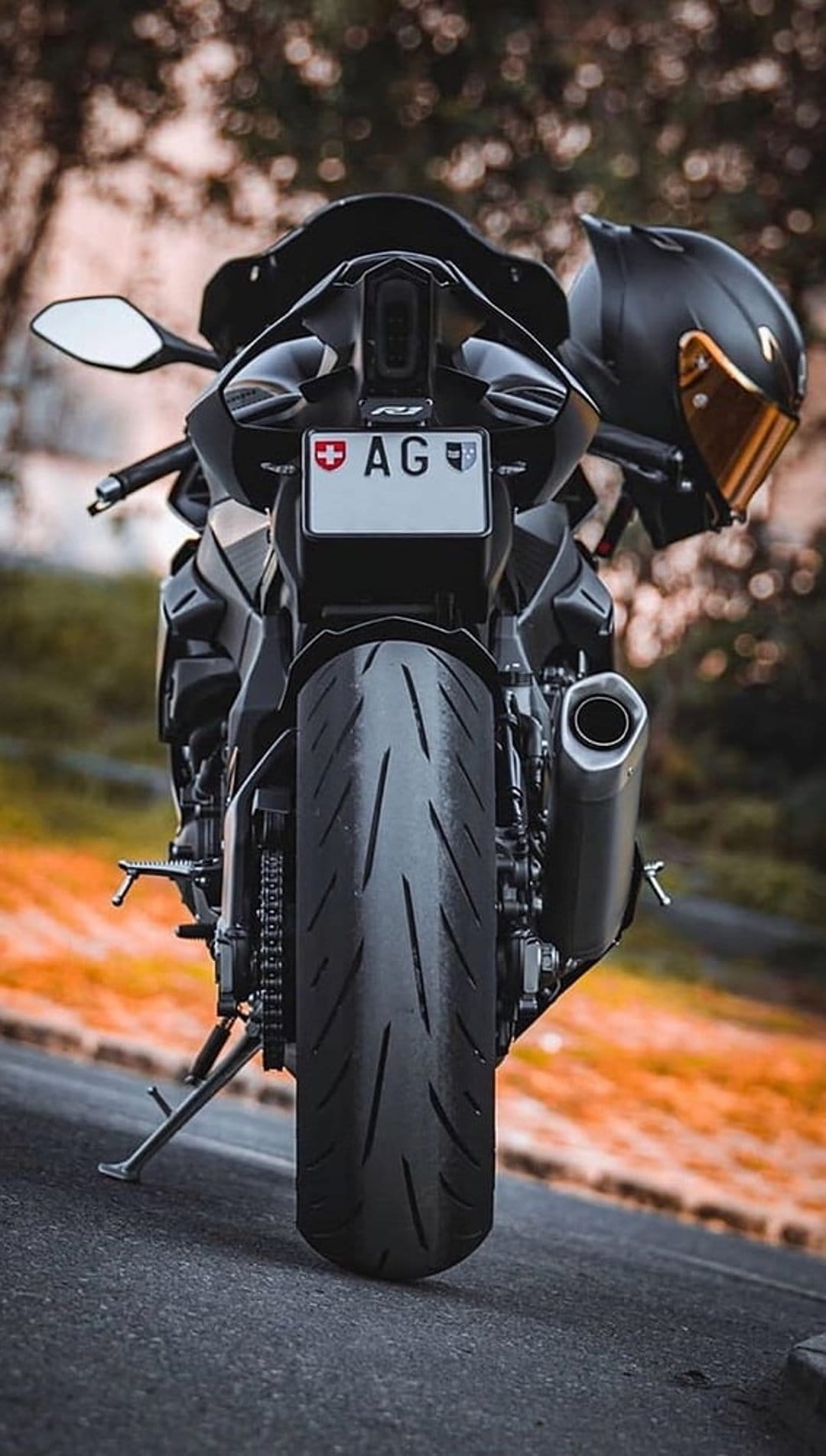Black sport motorcycle with a wide rear tire and a helmet with a golden visor parked against blurred autumn leaves.