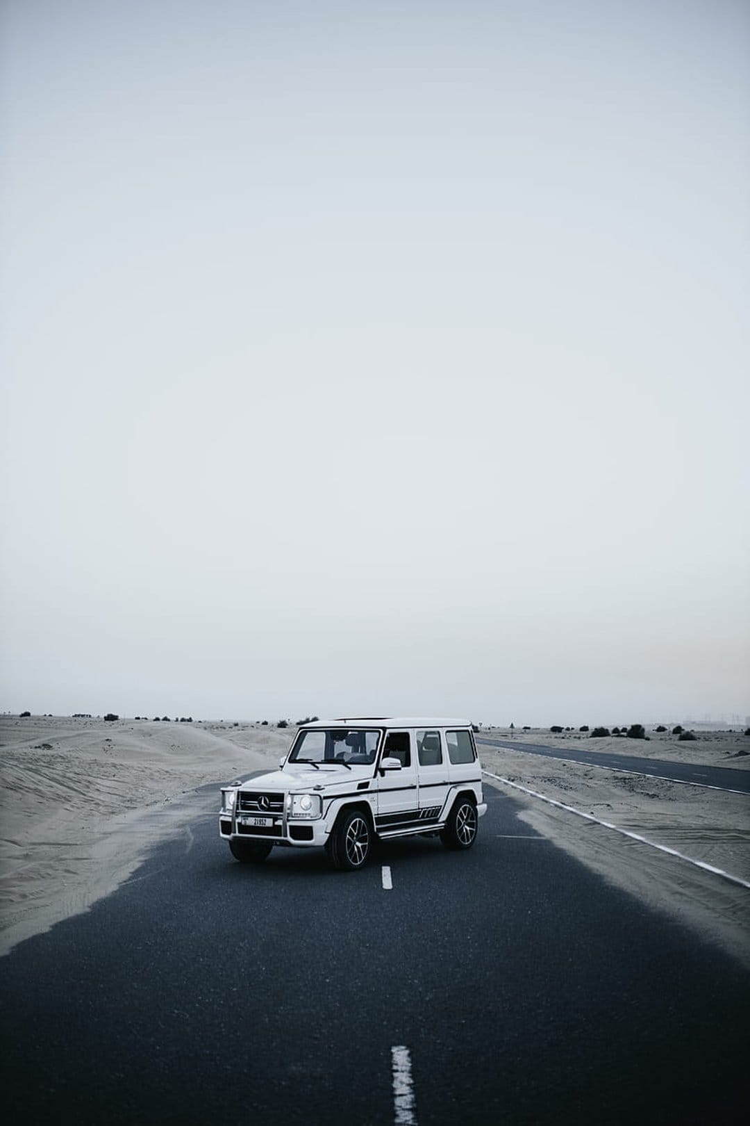 Mercedes G-Wagon driving on a straight desert highway under a pale overcast sky in a monochrome landscape scene.
