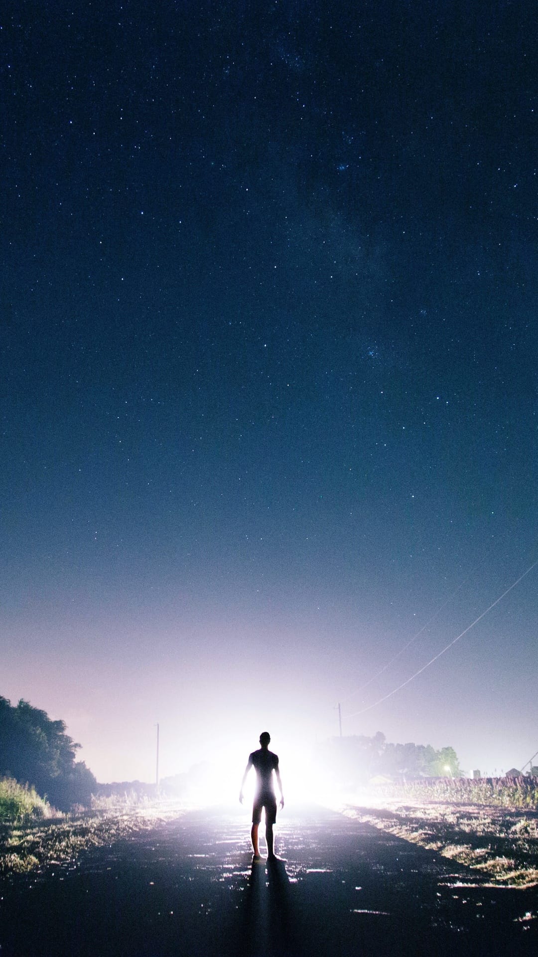 Lone person silhouetted by bright white light on a wet road at night under a clear starry sky with long shadows.
