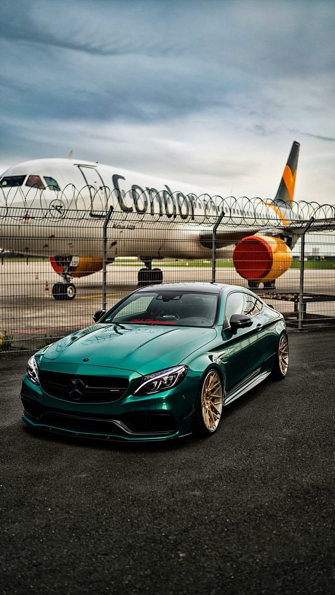 Emerald green Mercedes coupe with gold wheels parked on a runway in front of a striped Condor airplane.