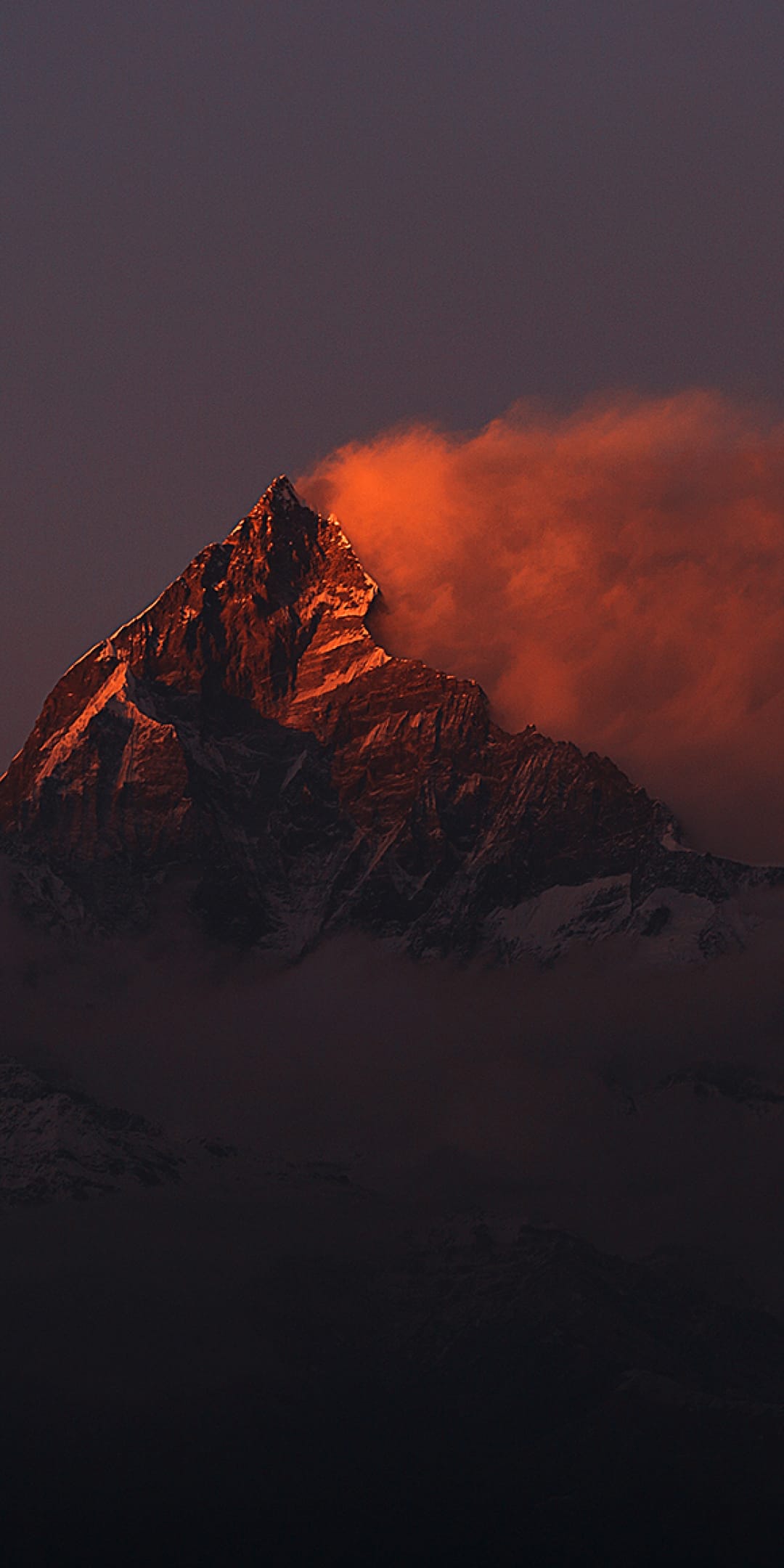Jagged mountain peak glowing orange under sunset light with dark misty slopes and swirling evening clouds.