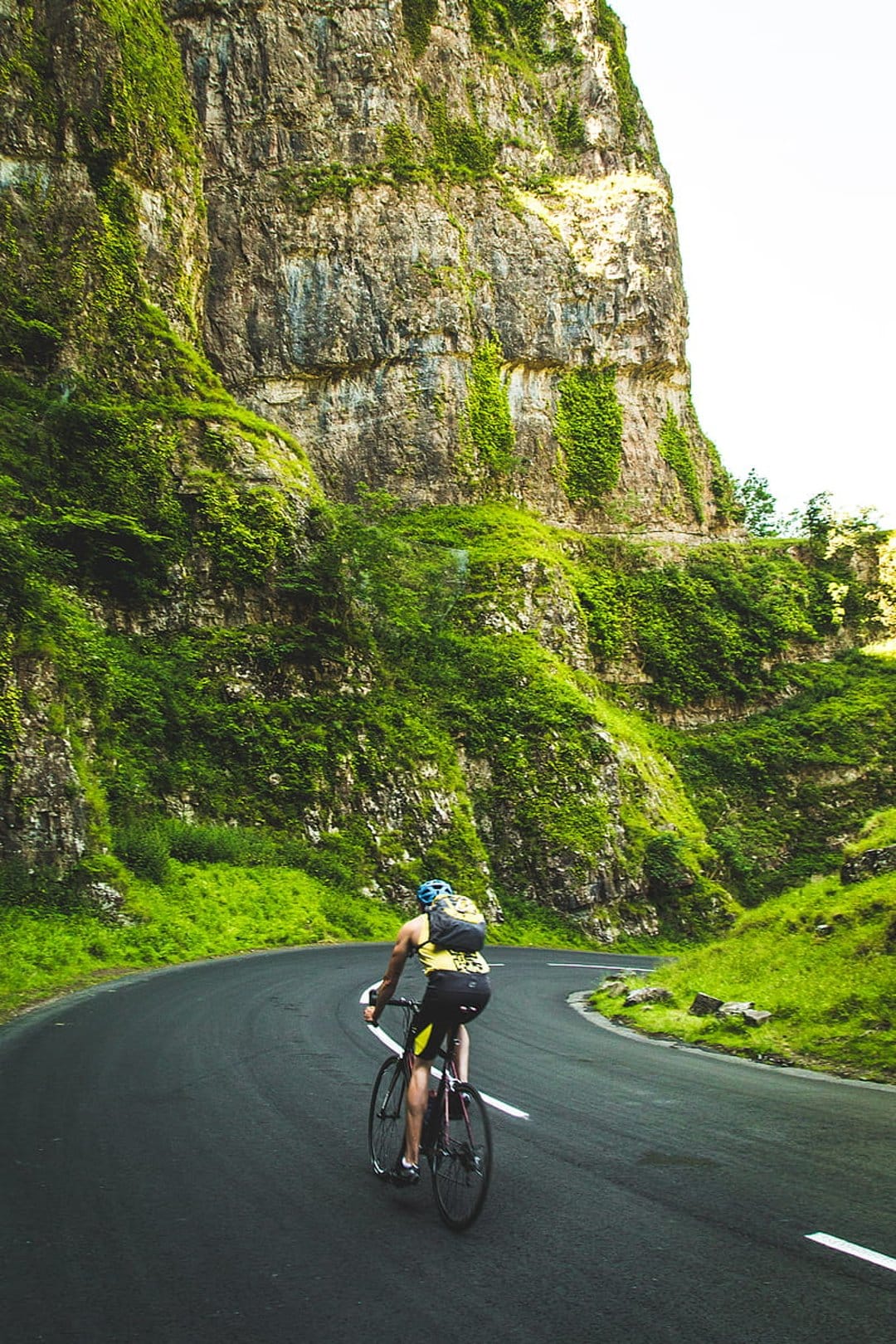 Cyclist on a winding asphalt road between towering mossy green cliffs and dense emerald vegetation.