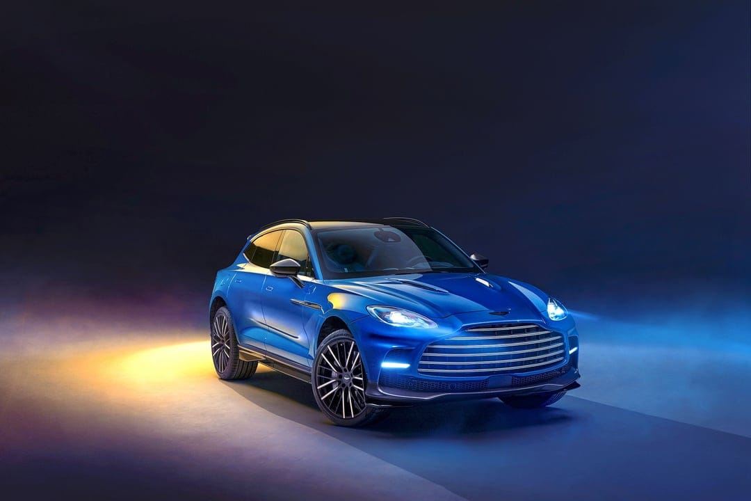 Blue Aston Martin DBX707 SUV with glowing headlights and multi-spoke wheels in a misty studio with yellow light.