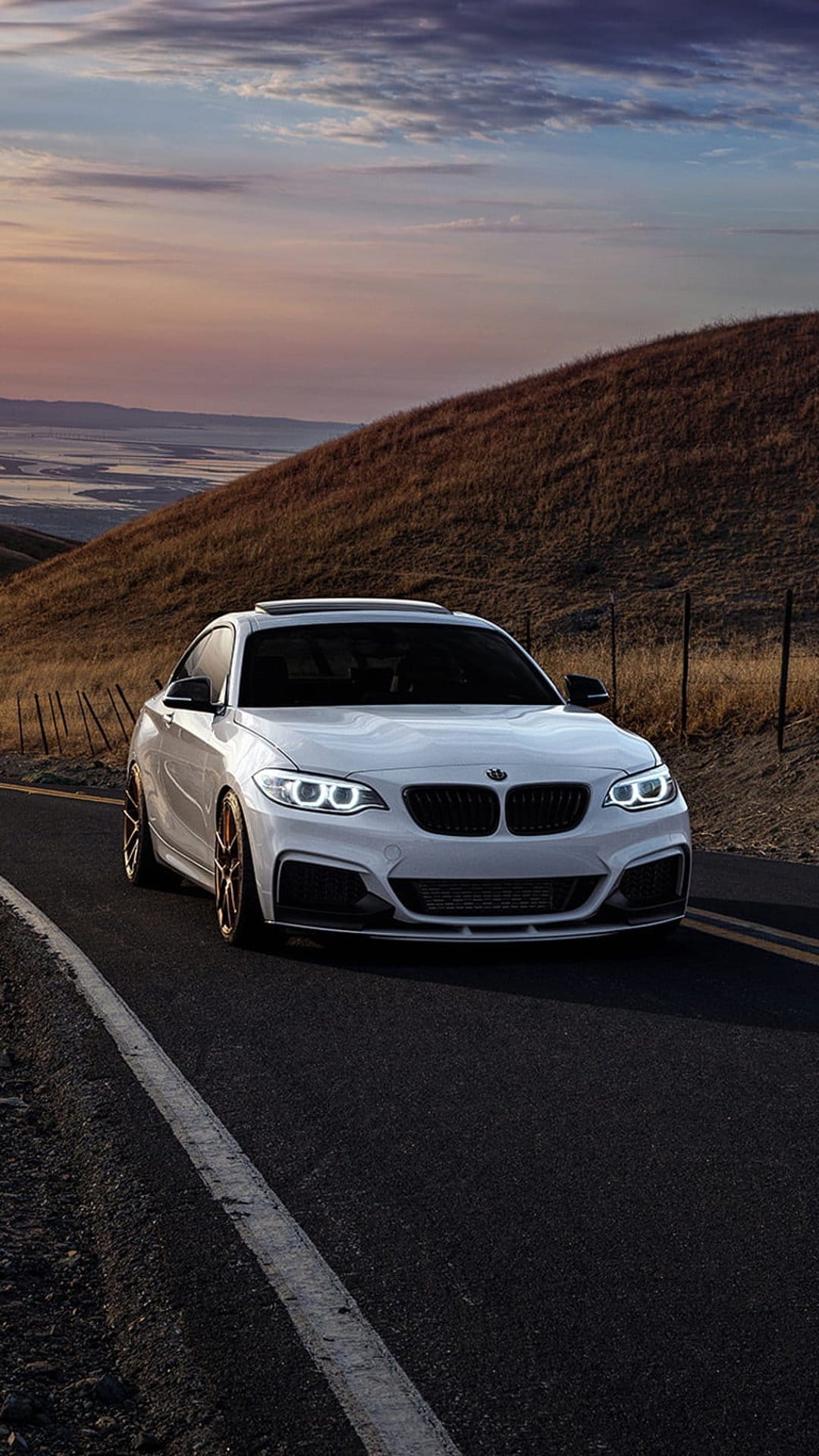 White BMW 2 Series Coupe with lit headlights cruises a winding mountain road under a pastel sunset sky.