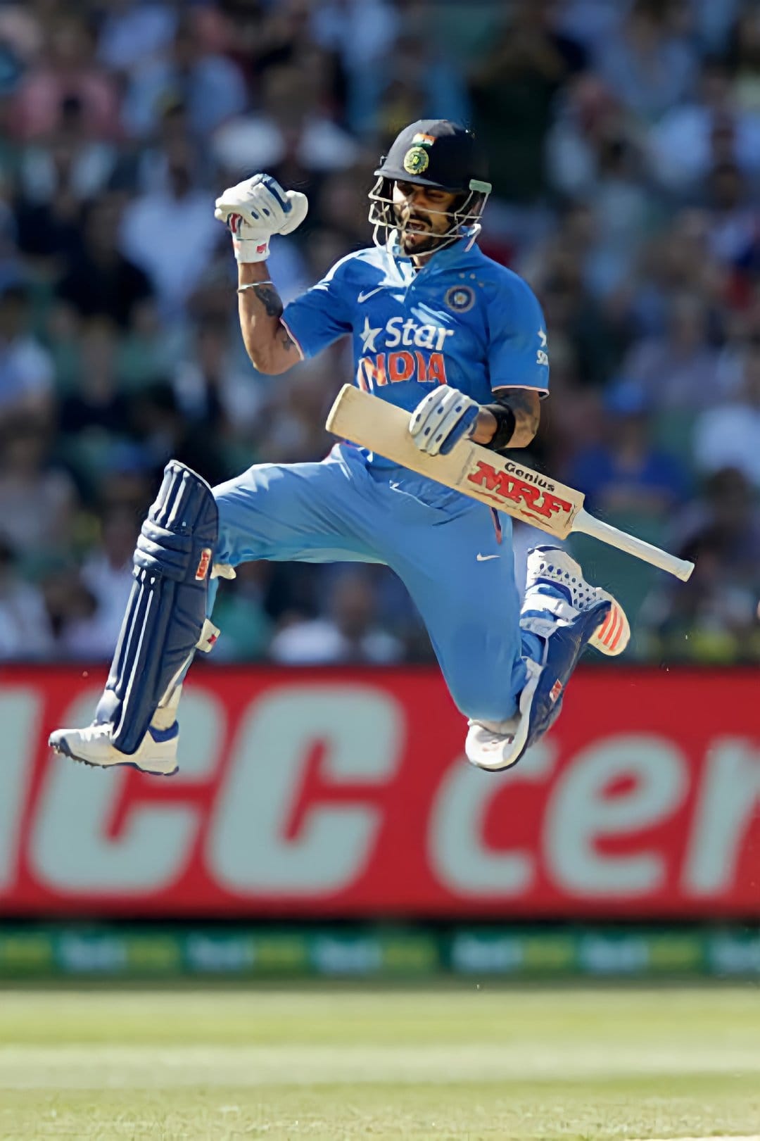 Virat Kohli leaps in celebration wearing a blue India jersey, helmet, and pads while holding a cricket bat.