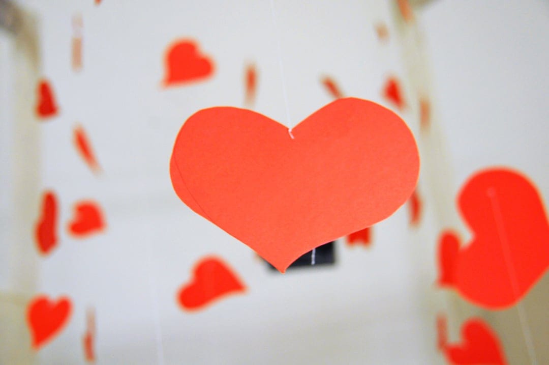 Red paper heart hangs from a mobile with blurred crimson hearts in a soft bokeh background for romance.