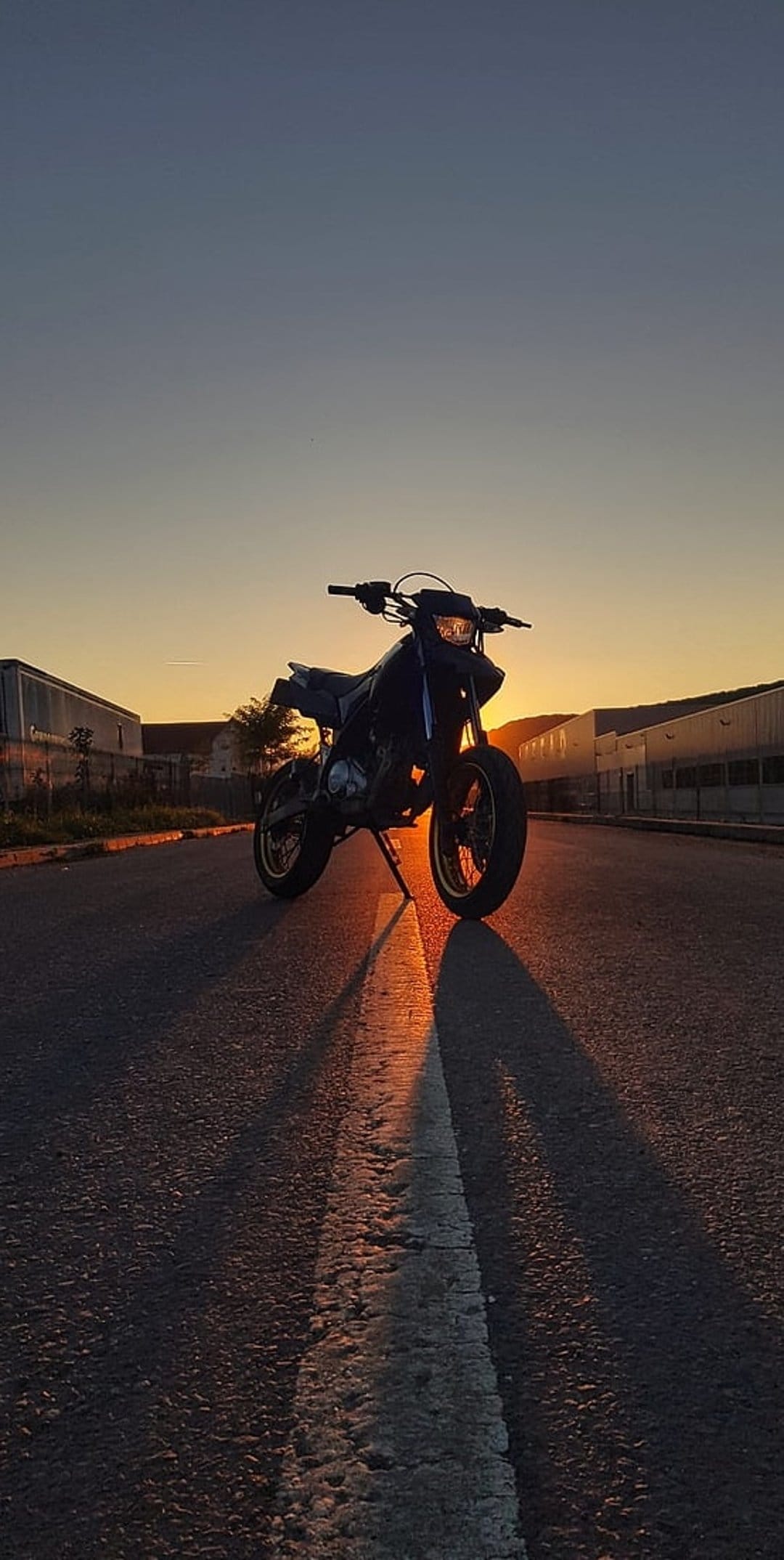 Motorbike parked on an asphalt road silhouetted against a vibrant orange sunset with industrial buildings behind.