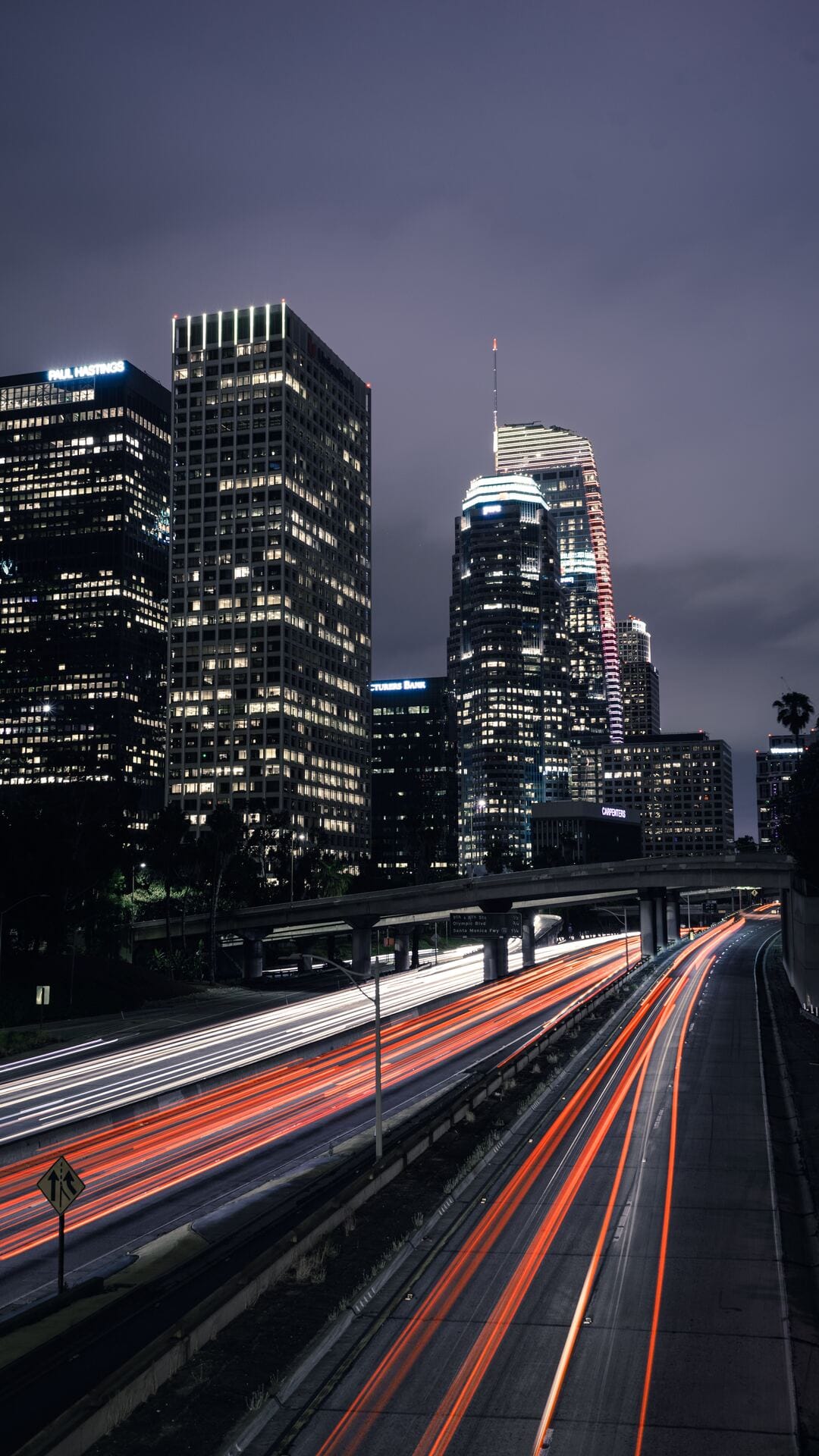 Red and white light streaks flow along a multi-lane highway surrounded by tall illuminated city buildings.