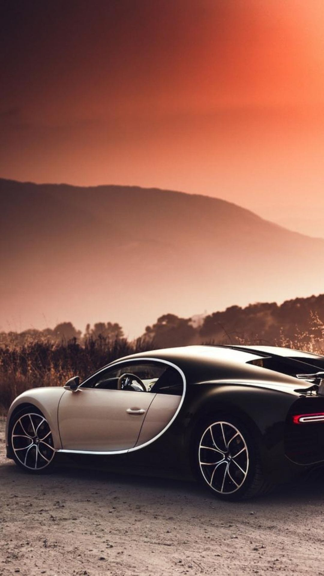 Bugatti Chiron hypercar parked on a dirt path against a glowing orange sunset sky and hazy mountain silhouettes.