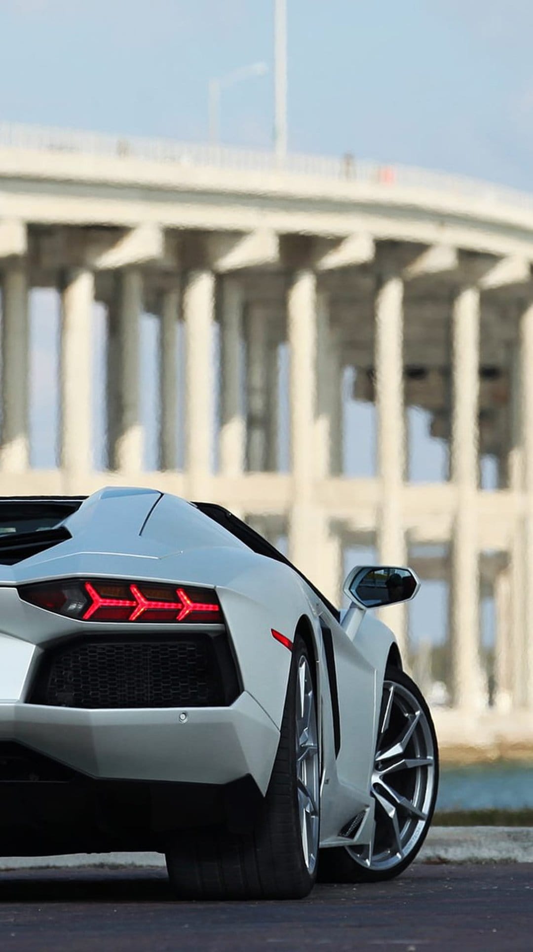 White Lamborghini Aventador Roadster rear view with red taillights parked under a massive concrete bridge pillar.