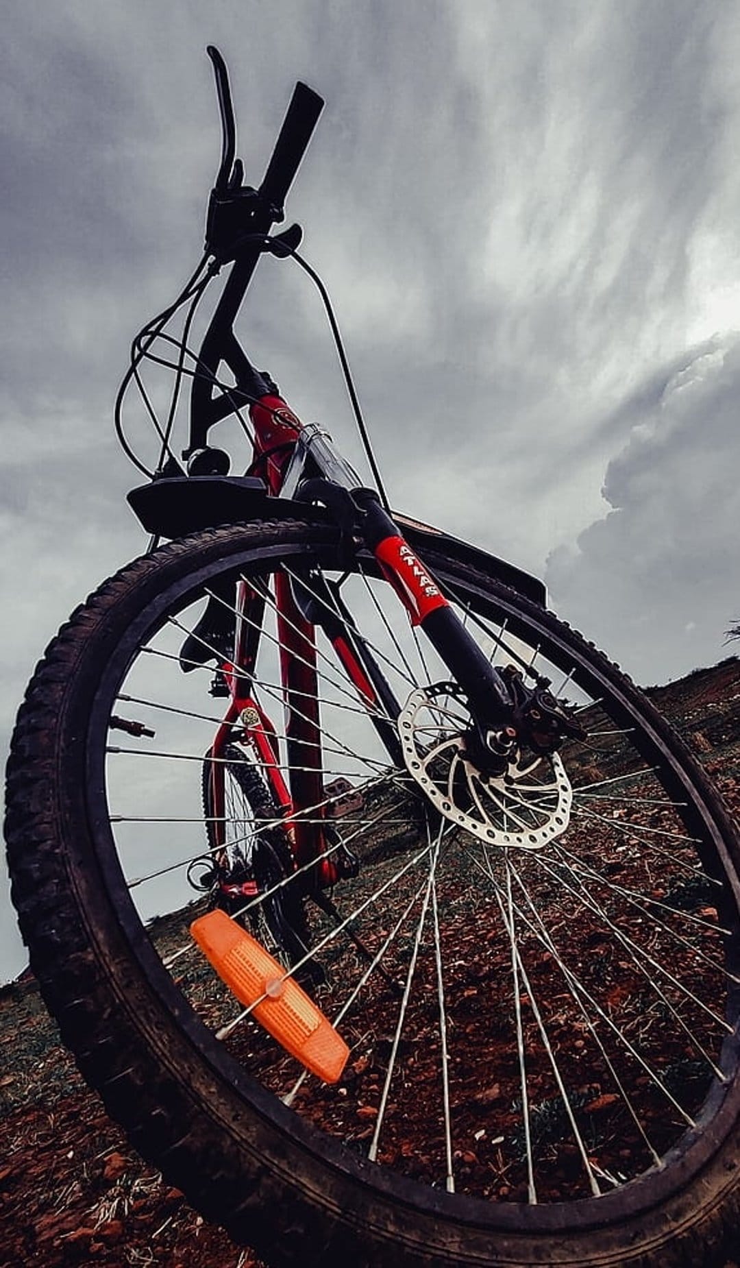 Red mountain bike front wheel and handlebars viewed from a low angle under a dark, dramatic cloudy sky on a trail.