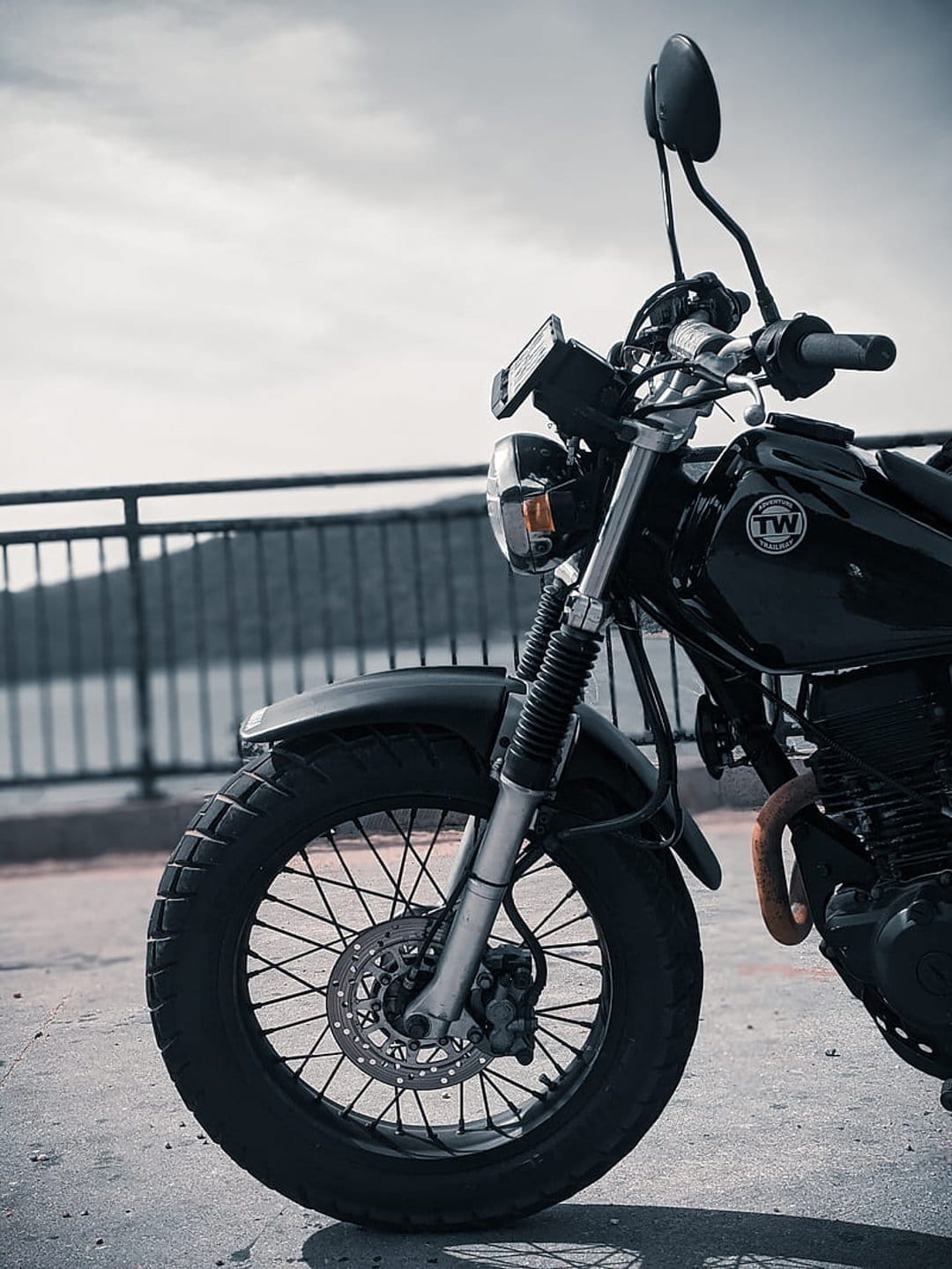 Black motorcycle front wheel and chrome headlight parked on concrete near a metal railing under a cloudy sky.