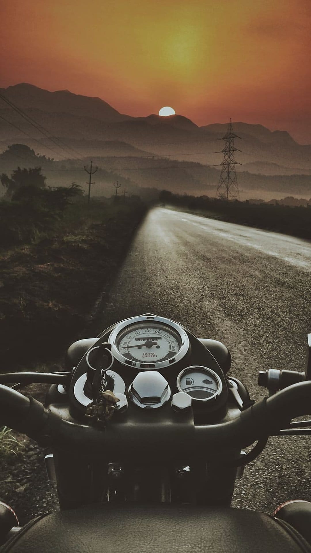 Motorcycle handlebars overlook a winding mountain road during a misty orange sunrise with power lines in view.