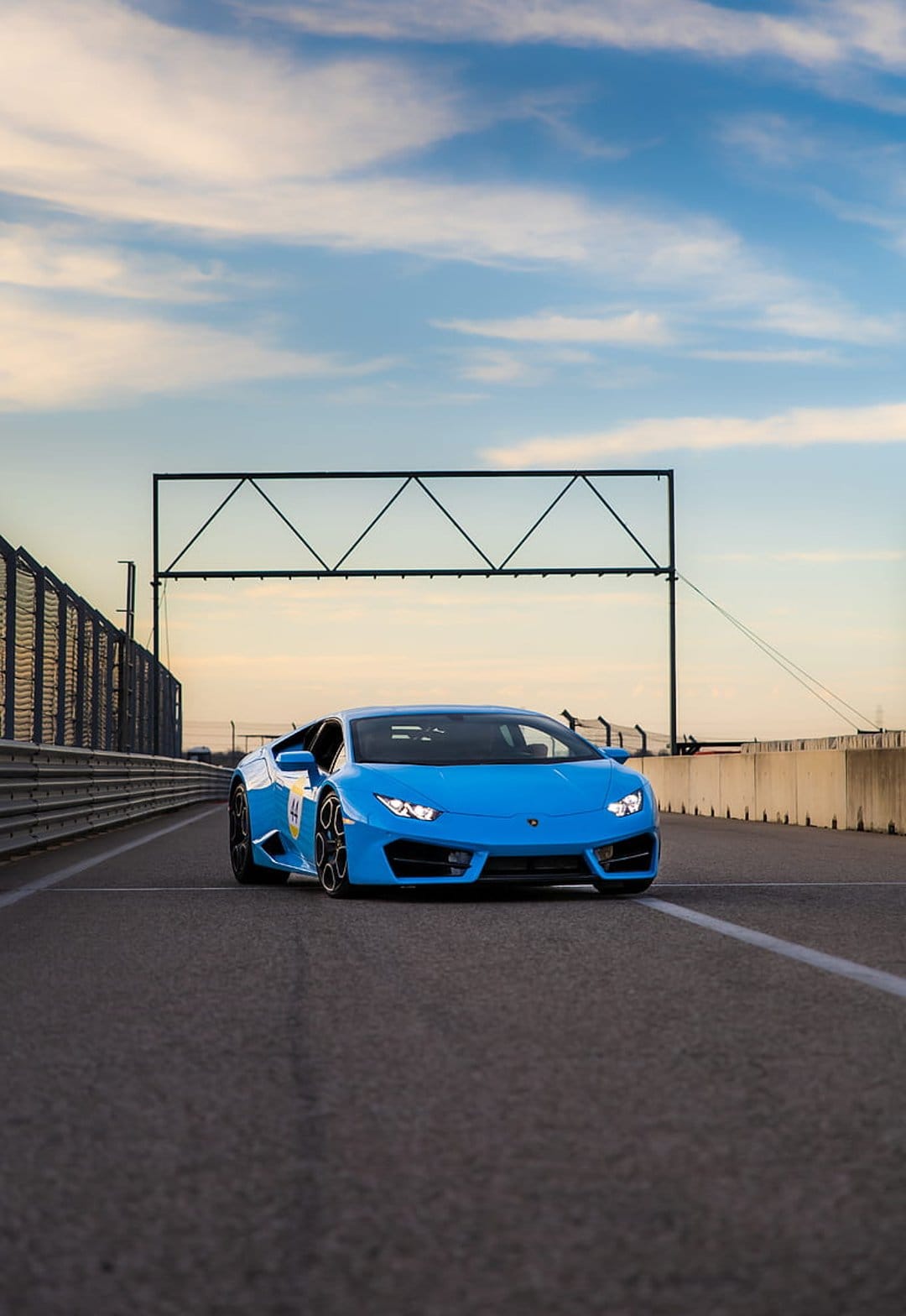 Blue Lamborghini supercar accelerates on a gray asphalt track under a cloudy sky near a metal stadium.