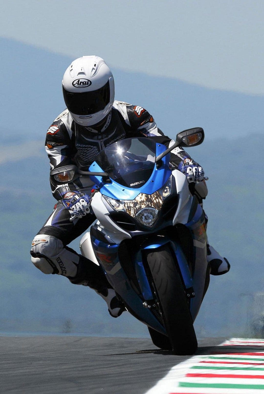 Motorcyclist in a black and white leather suit leans a blue Suzuki sportbike into a sharp, high-speed corner.