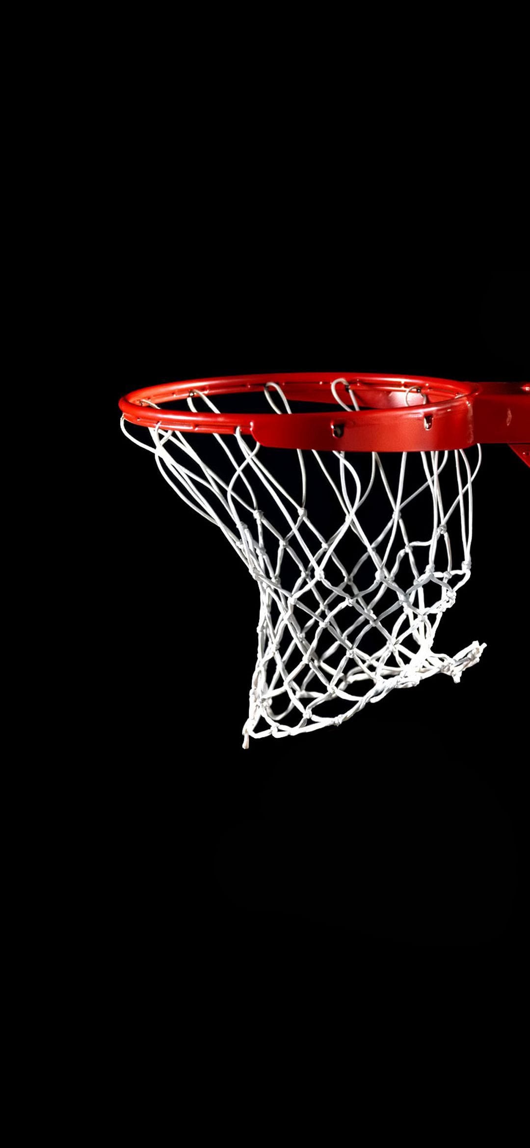 Red basketball rim with a white nylon net hangs against a solid black night sky in a close-up vertical shot.