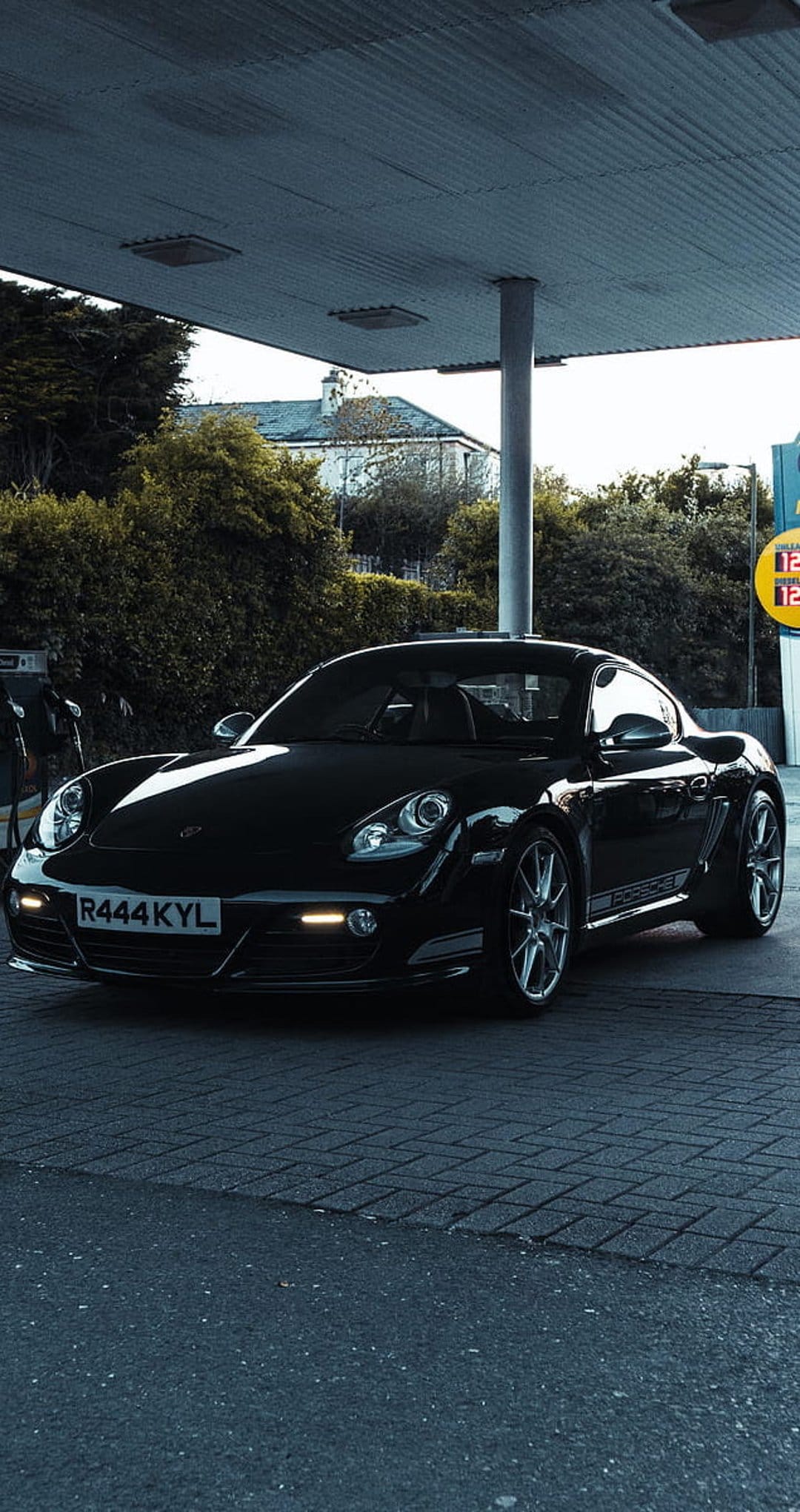 Black Porsche Cayman parked under a bright gas station canopy with headlights illuminating the ground at night.
