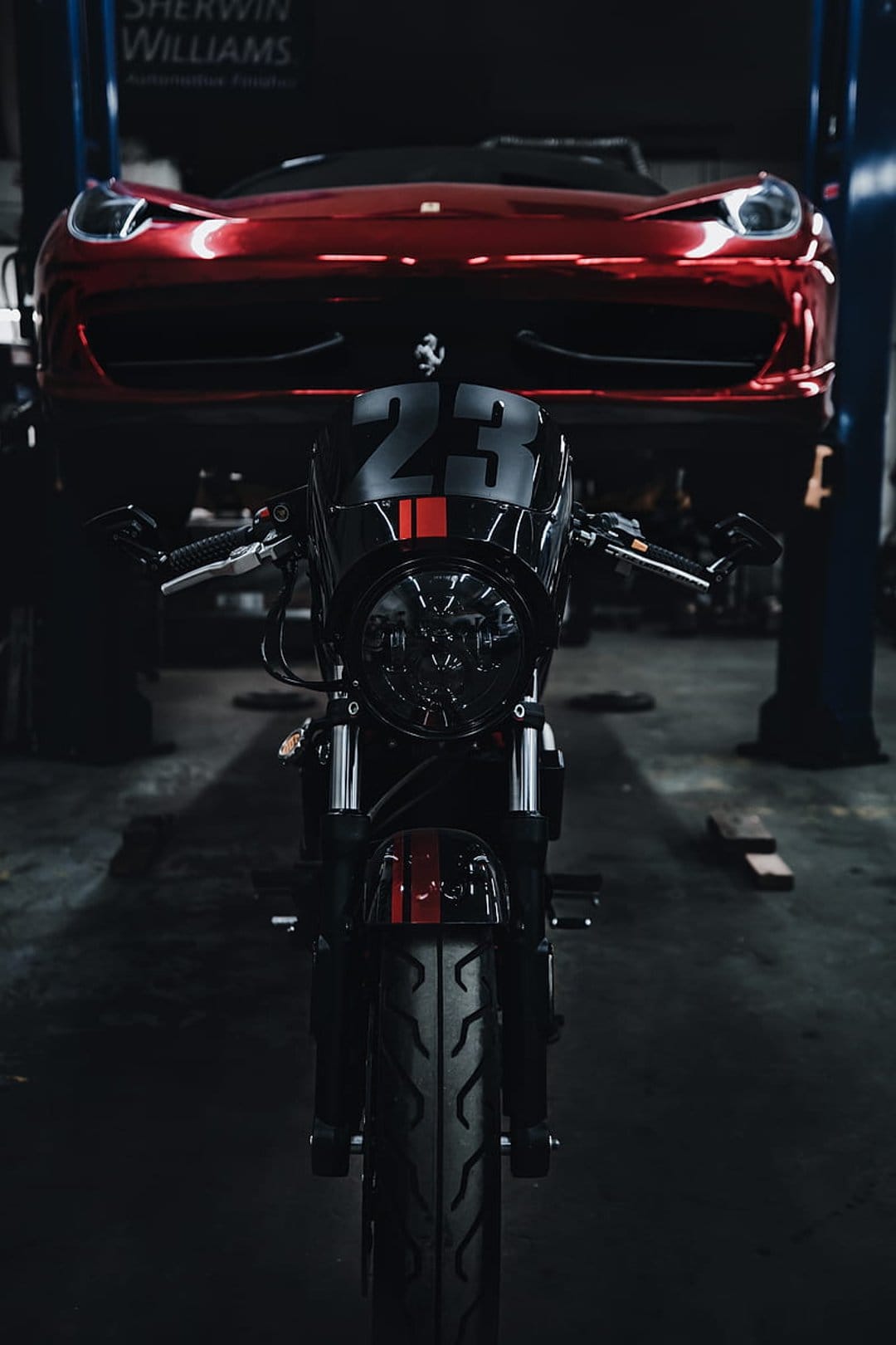 Black racing motorcycle number 23 parked in front of a red Ferrari on a car lift inside a dark workshop.