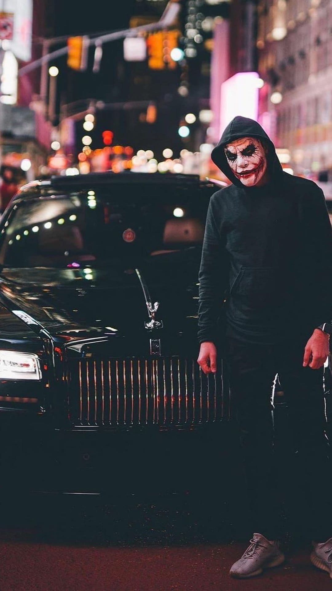 Man in Joker makeup and black hoodie stands by a sleek black sports car against a blurred neon city background.