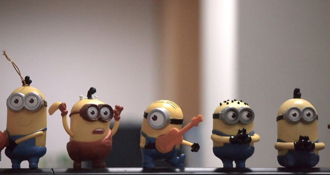 Five Minion toys with yellow skin and blue overalls pose on a shelf, including one character with a guitar.