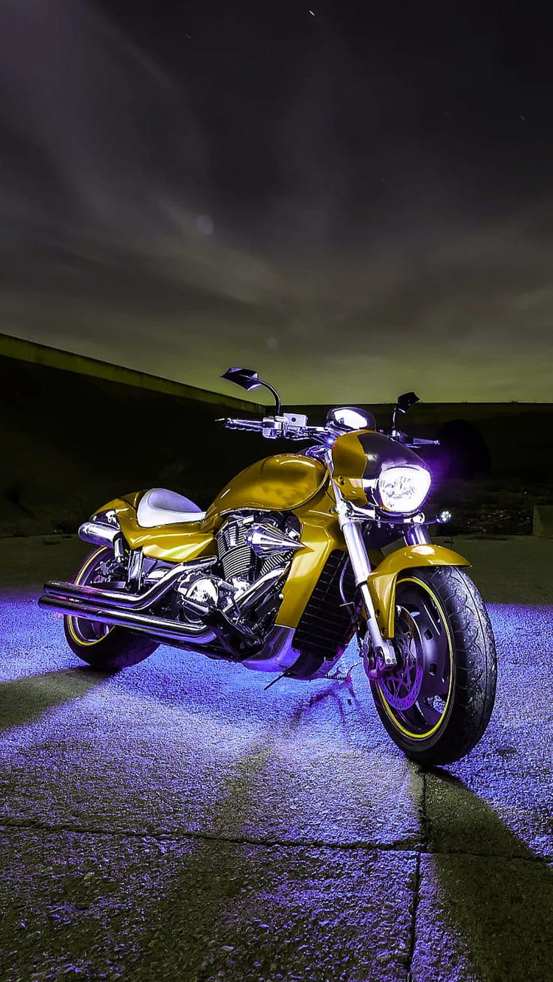 Gold motorcycle with vibrant purple underglow lighting parked on dark pavement under a cloudy night sky.