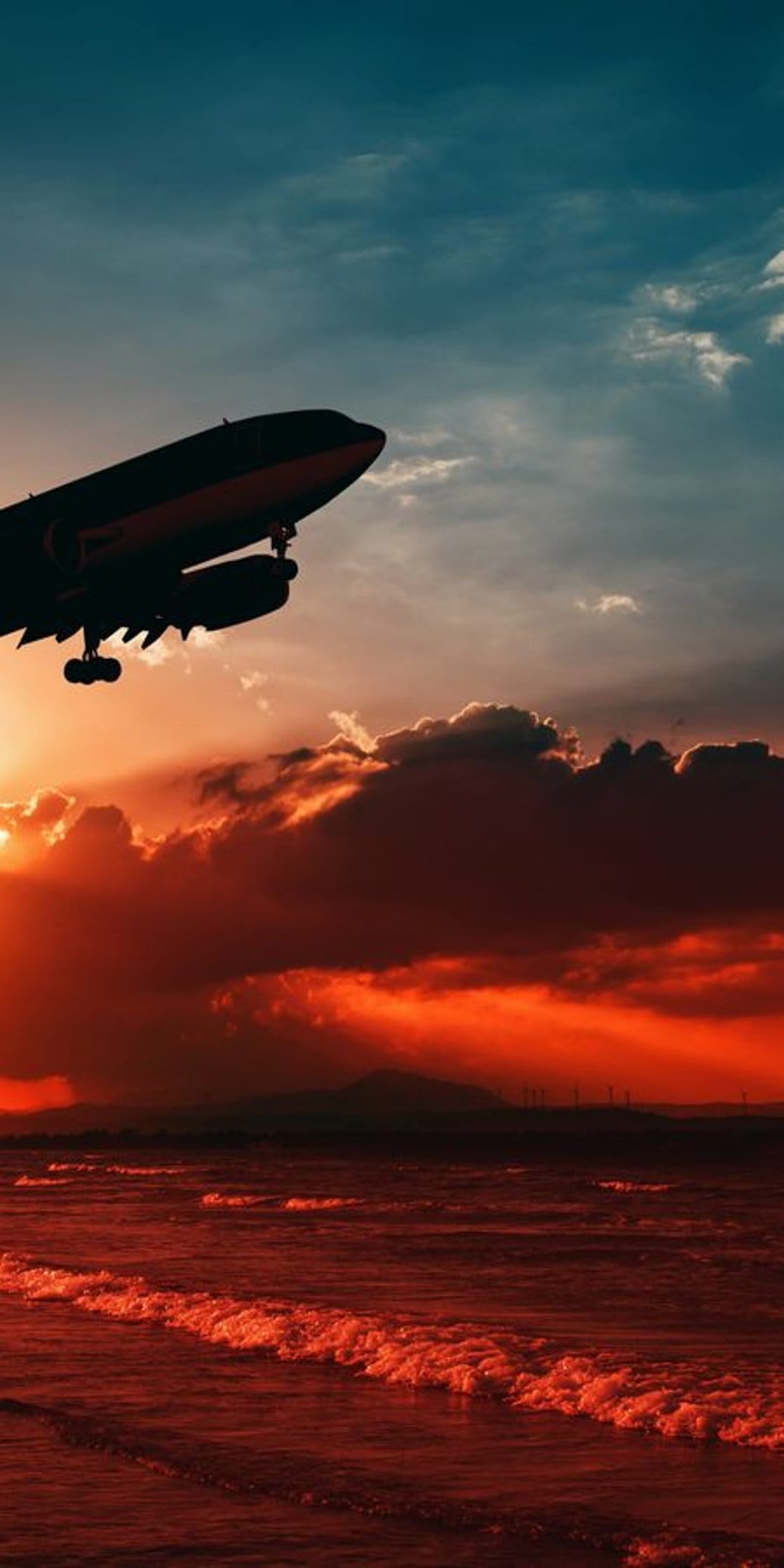 Passenger jet silhouette ascends over crashing ocean waves under a vibrant red and orange sunset sky.