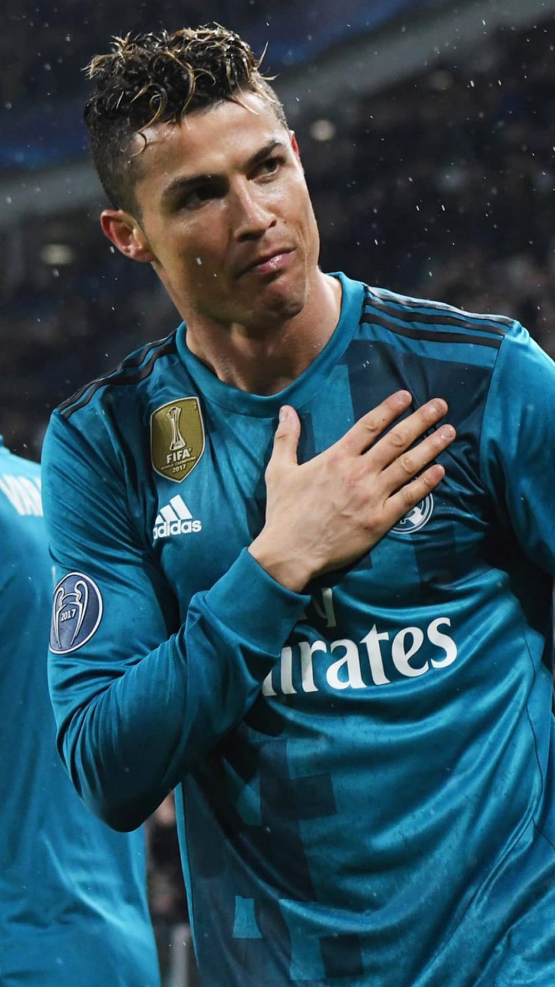 Cristiano Ronaldo rests his hand over his heart while wearing a teal jersey in a rainy football stadium.