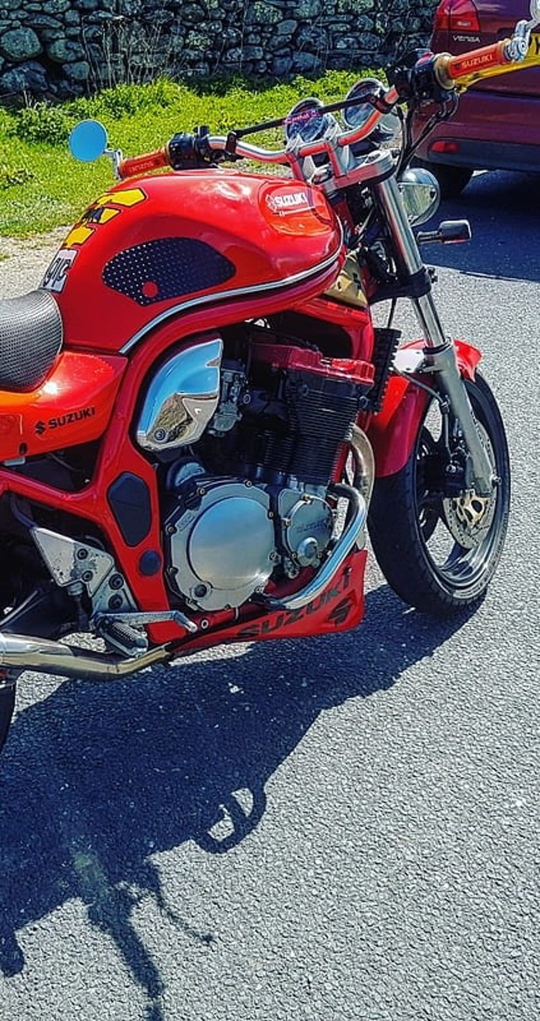 Suzuki motorcycle in red features a detailed engine and handlebars while parked on a sunlit asphalt road outdoors.
