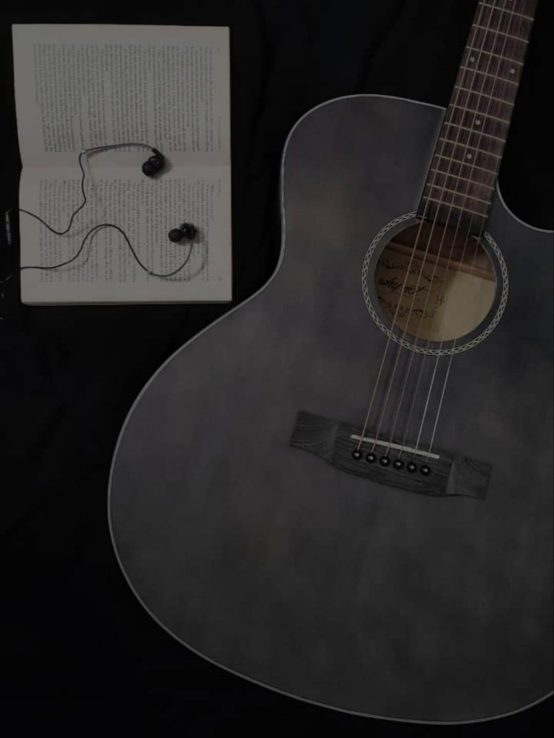 Acoustic guitar sits beside an open book with earbuds on a black surface in a vertical overhead view.