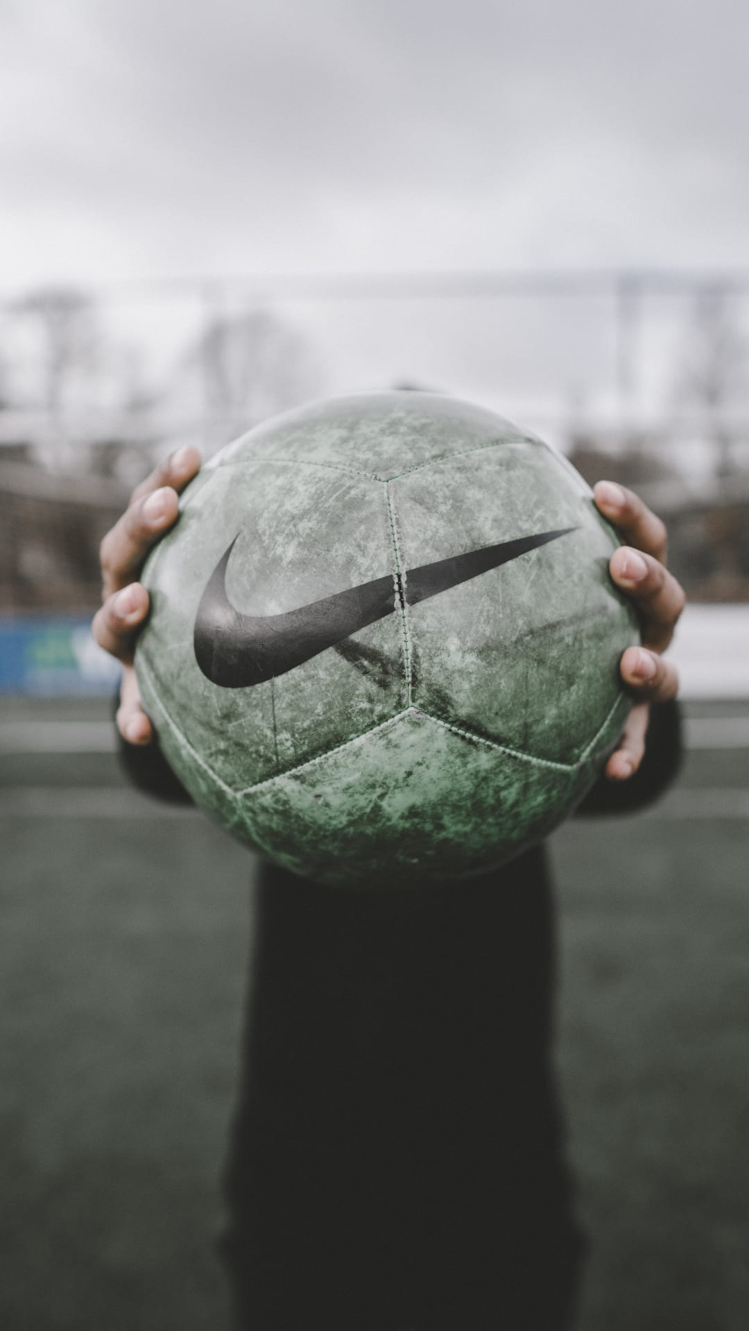 Hands hold a textured green Nike soccer ball with a black swoosh against a blurred outdoor sports field background.
