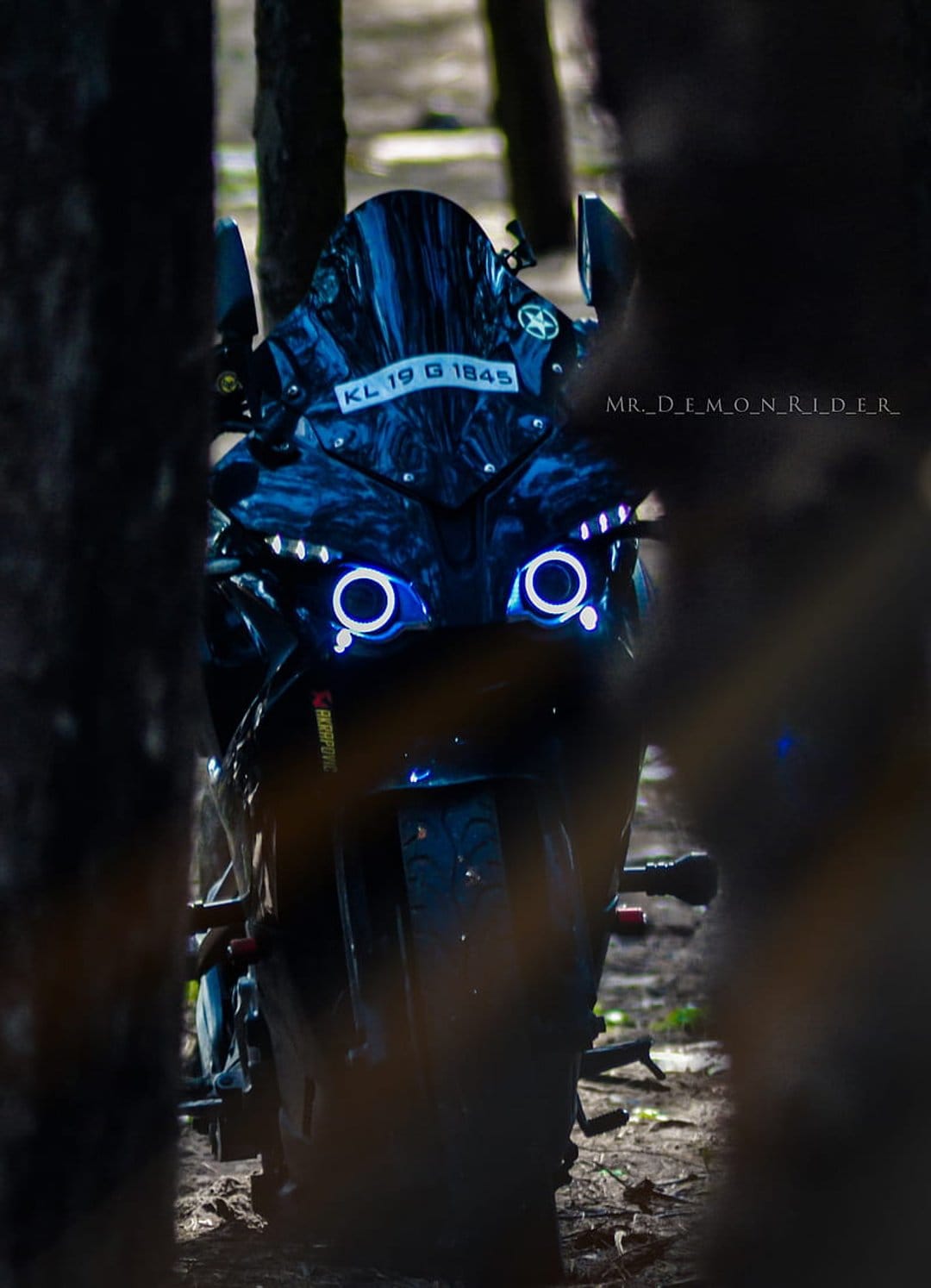 Black motorcycle with glowing blue circular headlights parked in a dark forest at night with visible license plate.