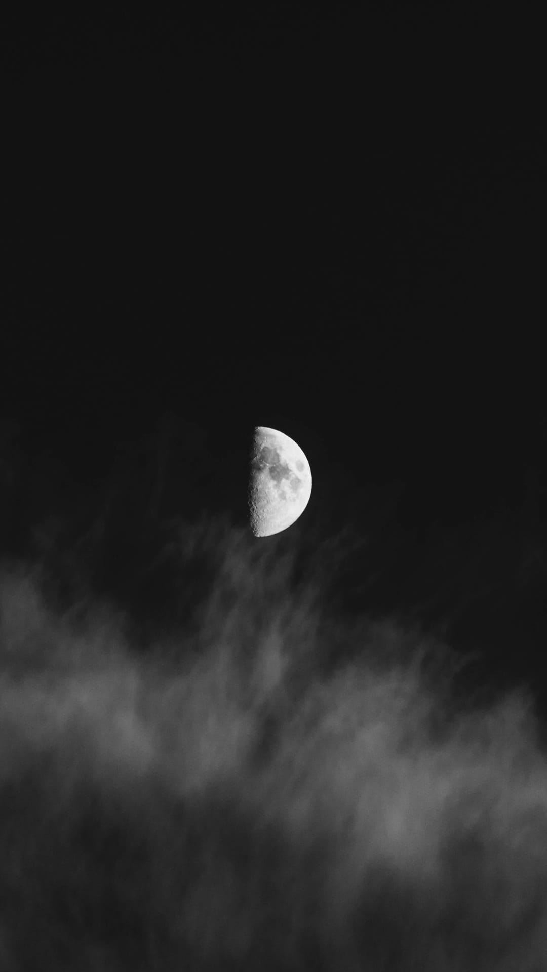Half moon with visible craters glowing above a layer of wispy, translucent clouds in a dark night sky.