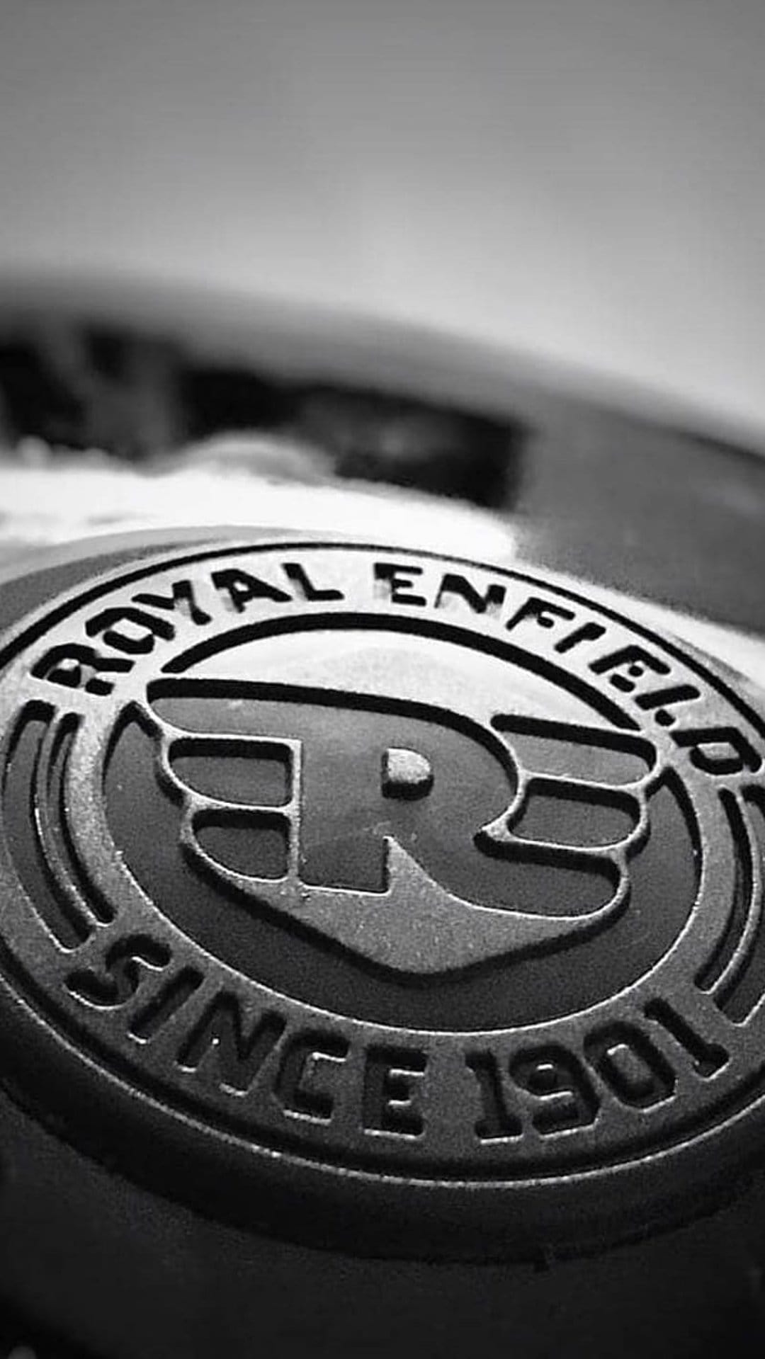 Royal Enfield logo 'R' emblem with circular text on a metallic surface in a black and white close-up shot.
