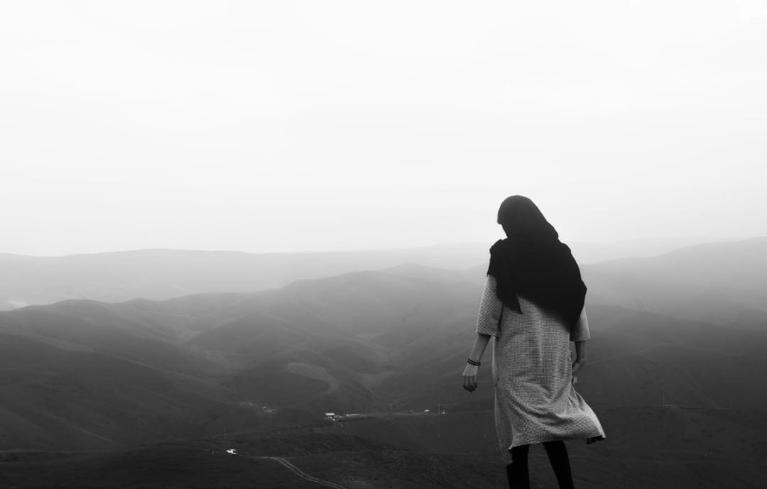 Woman wearing a hijab stands on a cliff edge facing distant misty mountains in a dramatic black and white scene.
