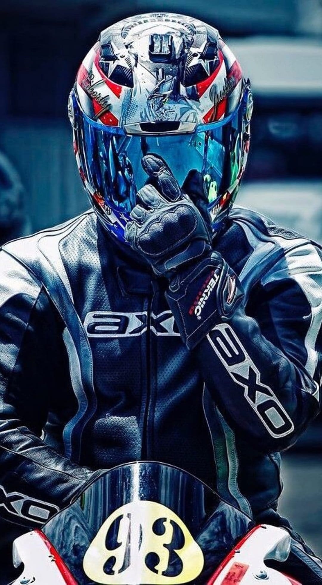 Motorcycle racer in a patriotic helmet and leather gear with the number 93 reflecting a blue sky in his visor.